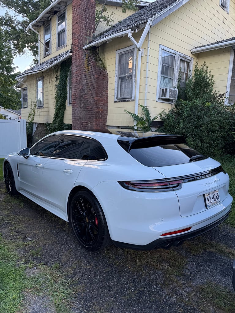 White Porsche Panamera parked in driveway next to yellow and brick house with green landscaping