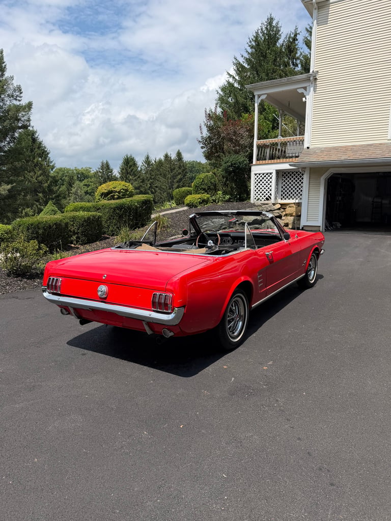 Red convertible Mustang parked in driveway next to house with landscaped yard and evergreen trees